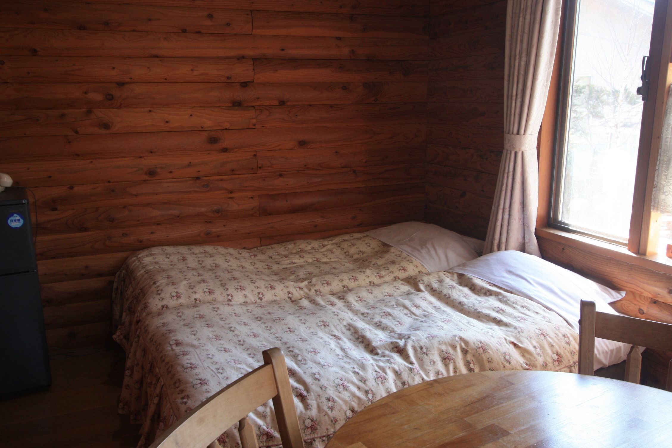 A wooden cabin room with a bed, a table, and a window with curtains.