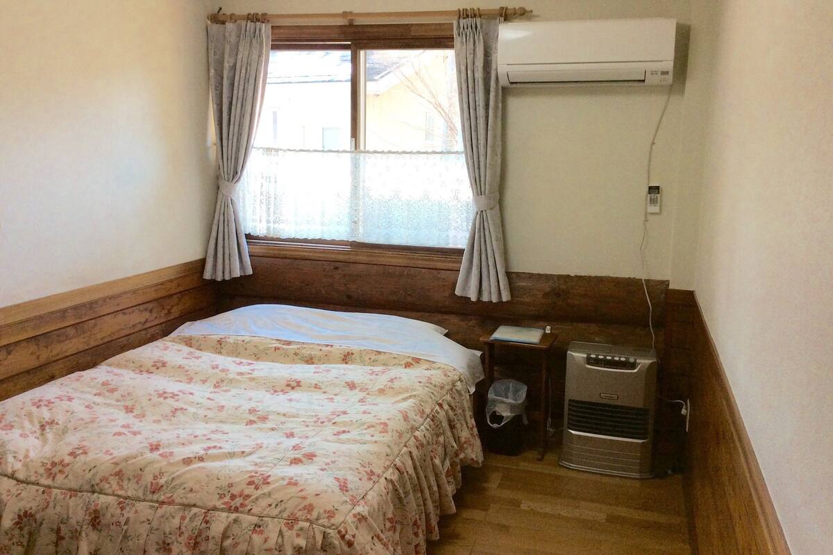 Ein Zimmer mit einem Bett, einem Vorhangfenster, einer Klimaanlage und einem kleinen Tisch mit Mülleimer.