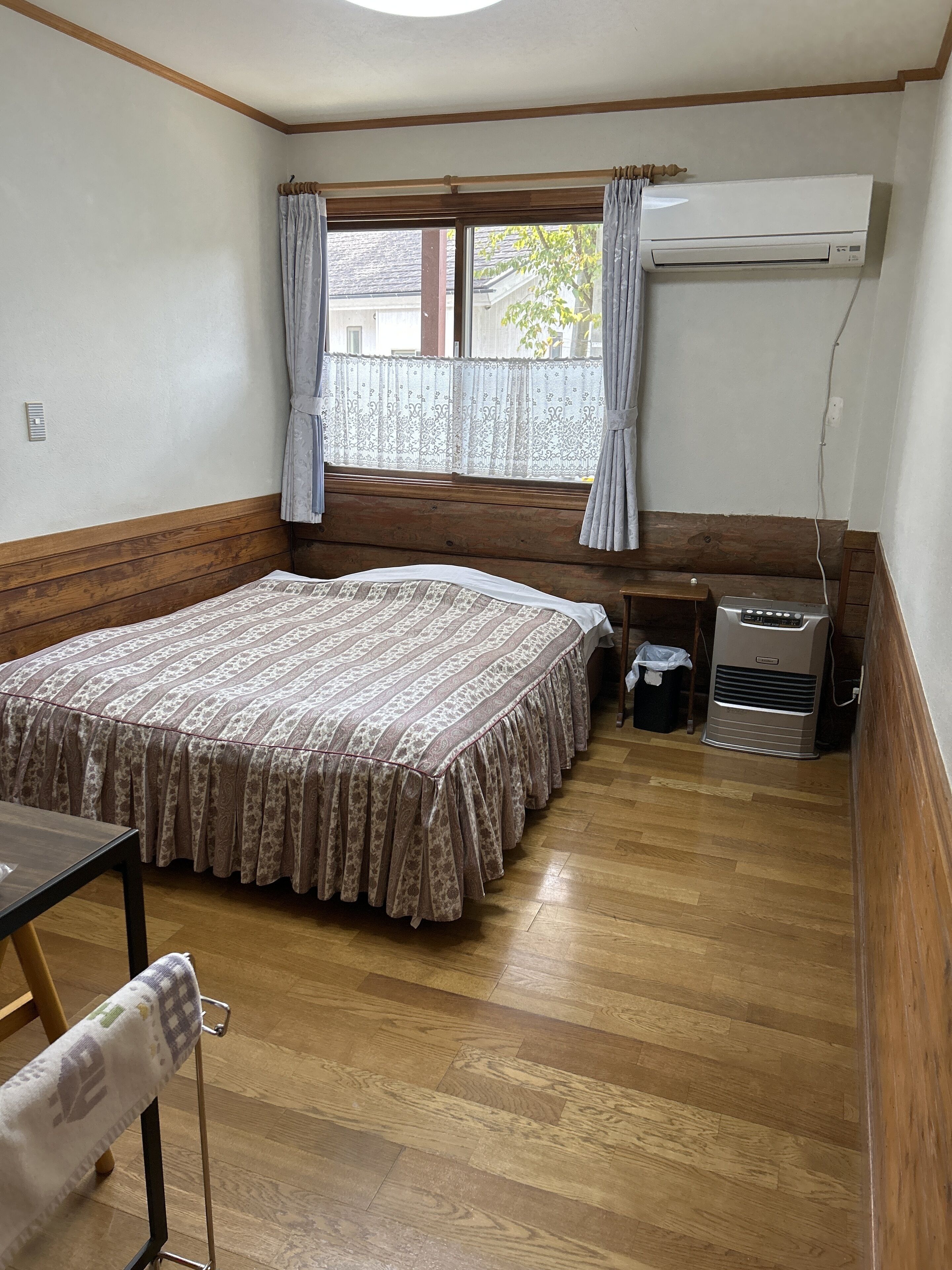 A room with a bed, a table, a chair, an air conditioning unit, and a window with curtains.