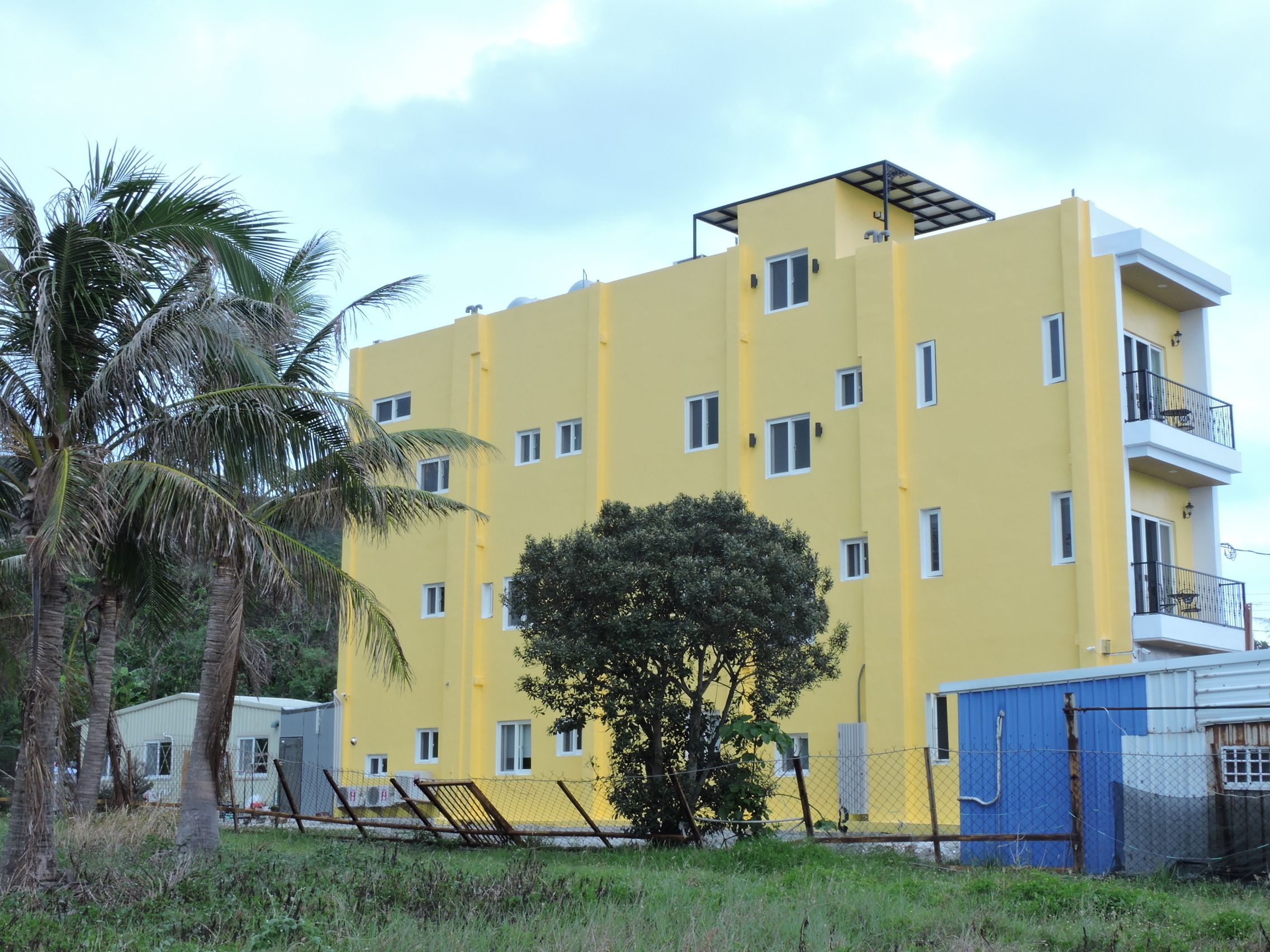 Un immeuble jaune à plusieurs étages, doté de nombreuses fenêtres et de balcons, entouré de palmiers et d’une zone délimitée par une clôture.