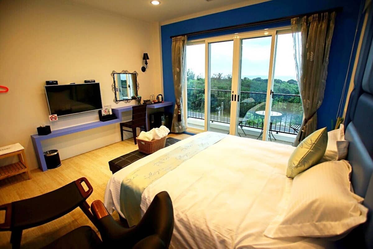 Habitación de hotel con una cama grande, televisión, escritorio con silla, balcón con vistas y ventana con cortinas.