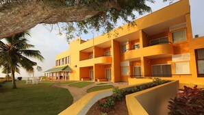 Un edificio de varios pisos con balcones, un porche cubierto y un jardín ajardinado con palmeras y arbustos.