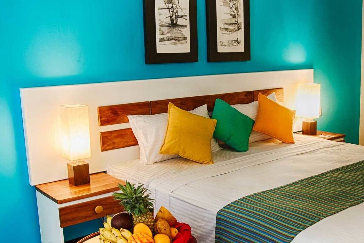 A hotel room with a bed, colorful pillows, a bedside table with a lamp, and a fruit basket.