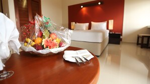 Una habitación de hotel con una cama bien hecha, una mesita de noche con una cesta de frutas y una servilleta blanca, y un tocador de madera con una lámpara.
