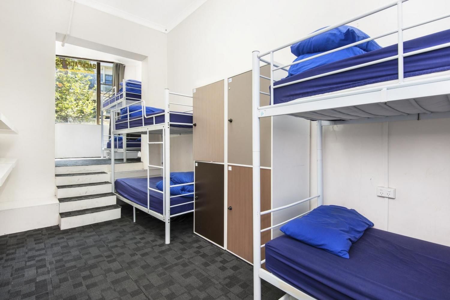 A dormitory room with bunk beds, a staircase, and a window with a view of greenery.