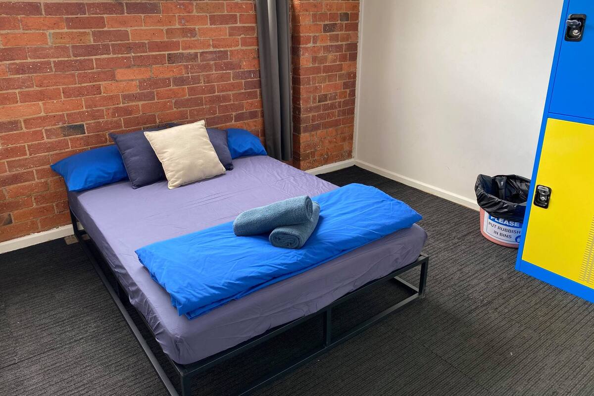 A single bed with a metal frame, a purple mattress, and blue bedding. A grey towel is placed on the bed. The room has a brick wall and a blue door with a yellow panel. A trash bin is visible on the floor.