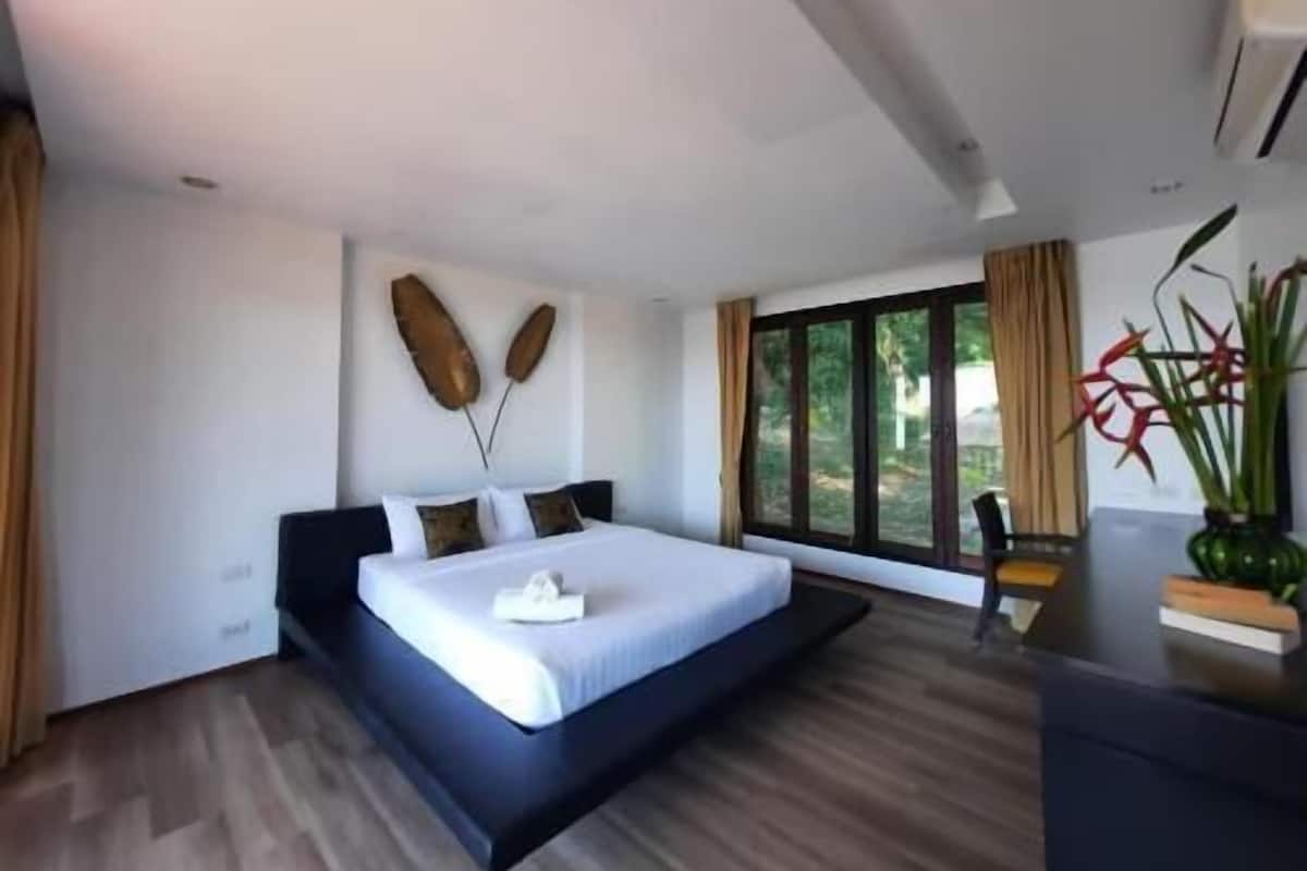 A modern bedroom with a large bed, a wall-mounted artwork, a window with curtains, and a desk with a plant.