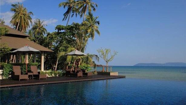 Zona junto a la piscina con sillones reclinables y sombrillas, con vista al mar tranquilo y palmeras.
