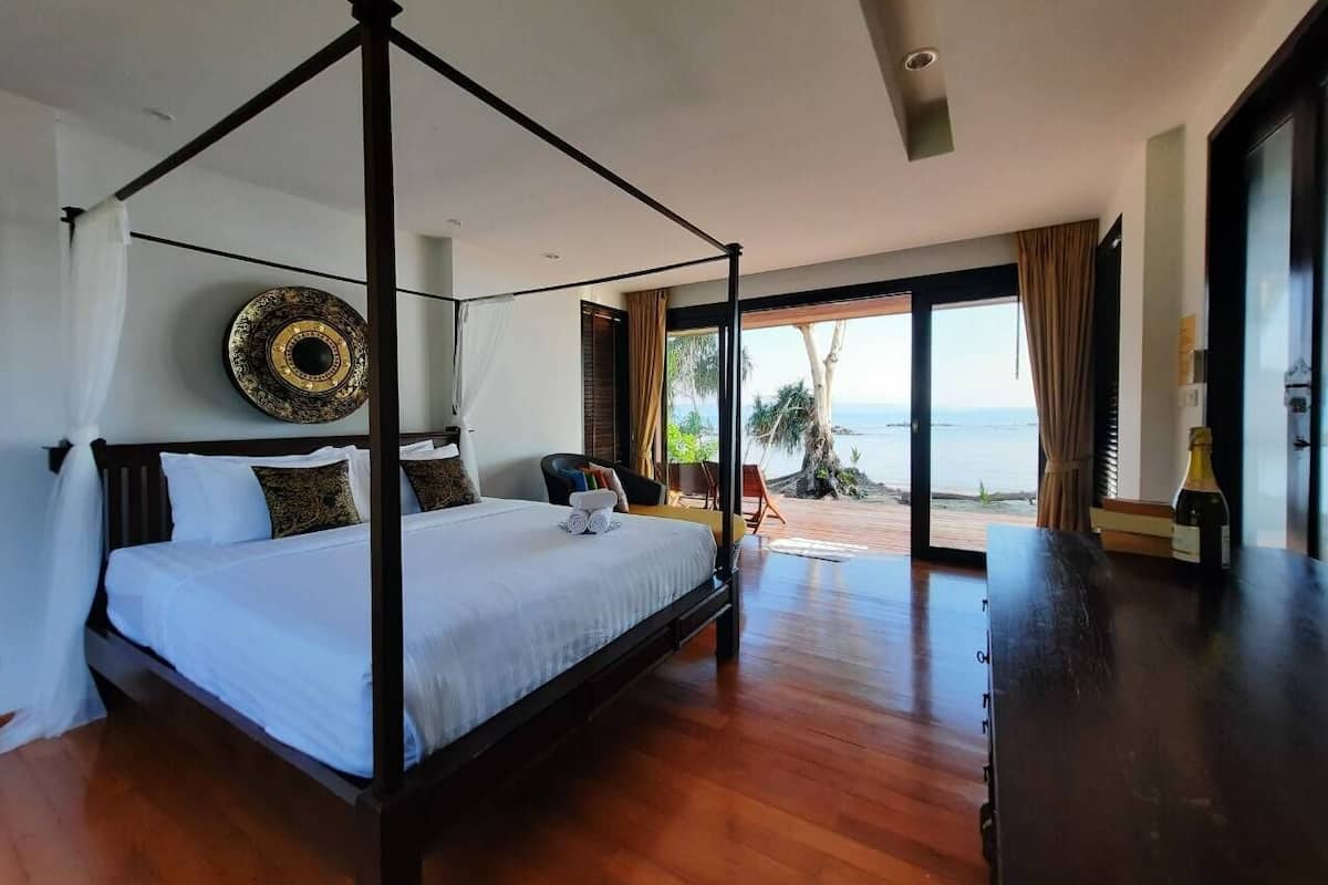 A four-poster bed with a white bedspread, a wooden floor, a view of the ocean, and a large decorative mirror on the wall.