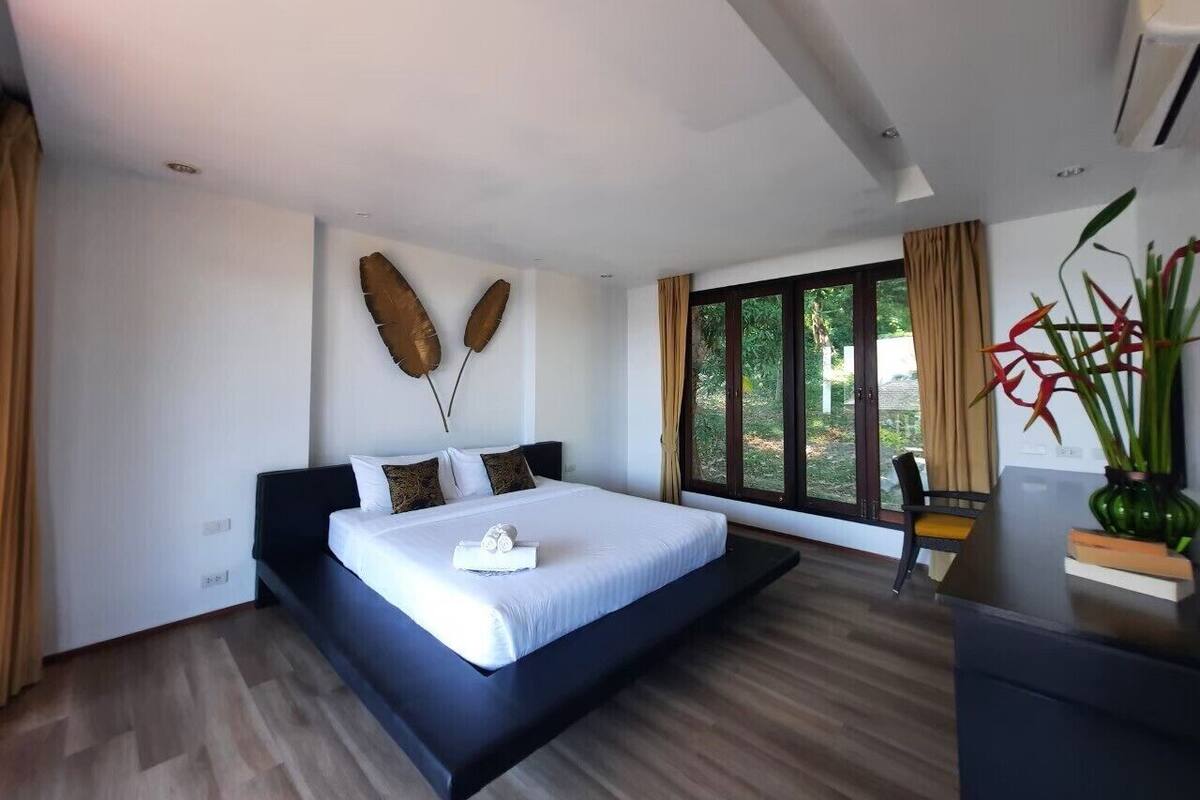 A modern bedroom with a large bed, a desk, and a view of greenery outside.