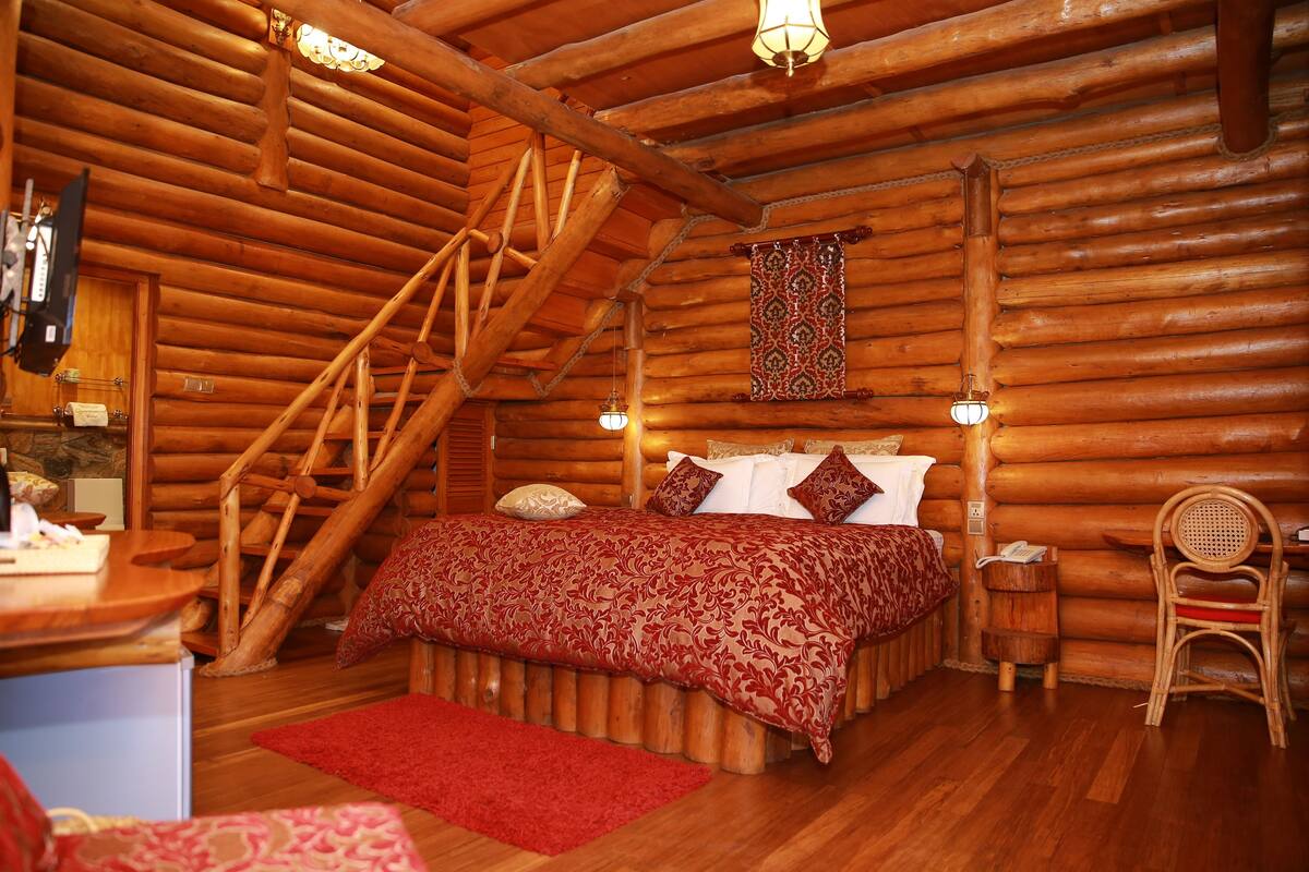 Un dormitorio de cabaña con escalera de madera, una cama con ropa de cama estampada, una mesita de noche de madera y una silla.