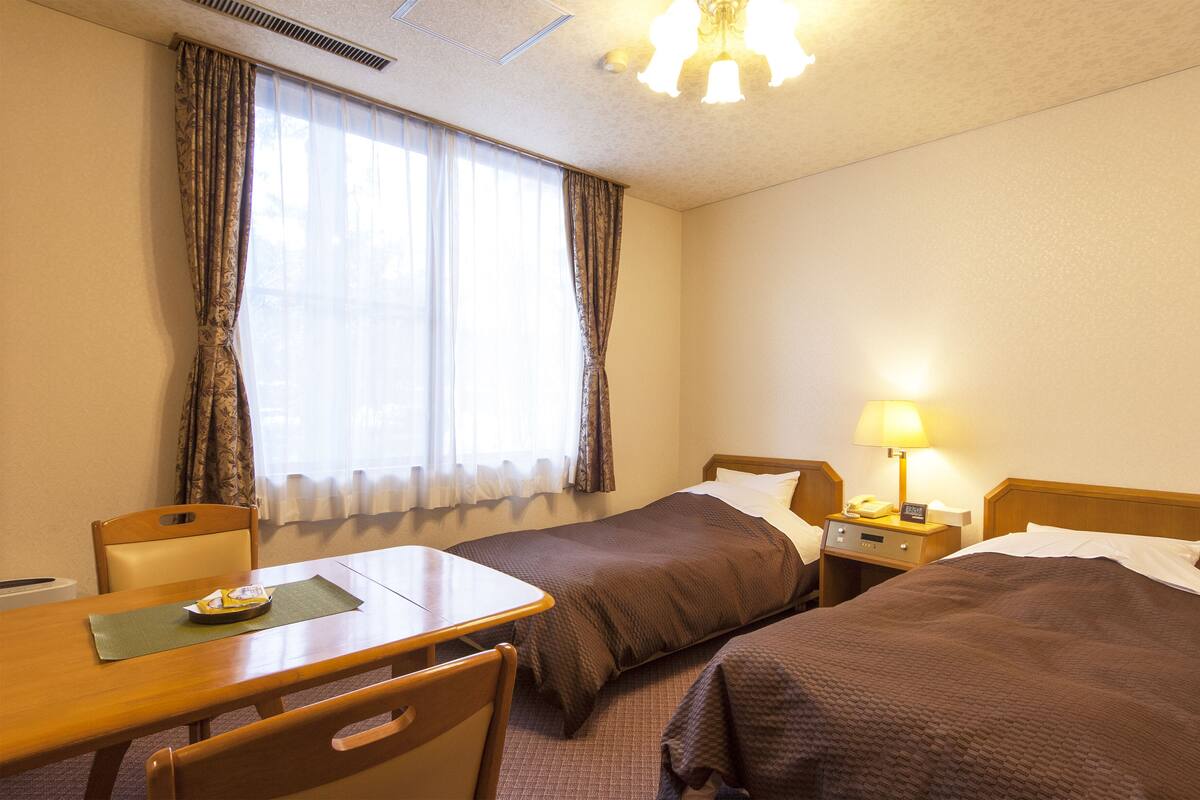 Ein Hotelzimmer mit zwei Einzelbetten, einem Holztisch mit Stuhl, einem Schreibtisch, einer Lampe und einem Fenster mit Vorhängen.
