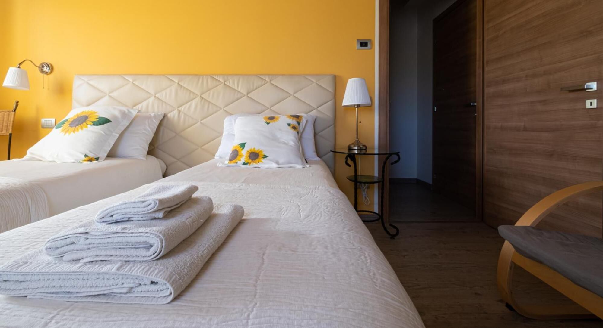 A hotel room with two beds, each with a headboard, and a wooden bench. There are folded towels on the beds, a lamp on a table, and a door leading to another room.