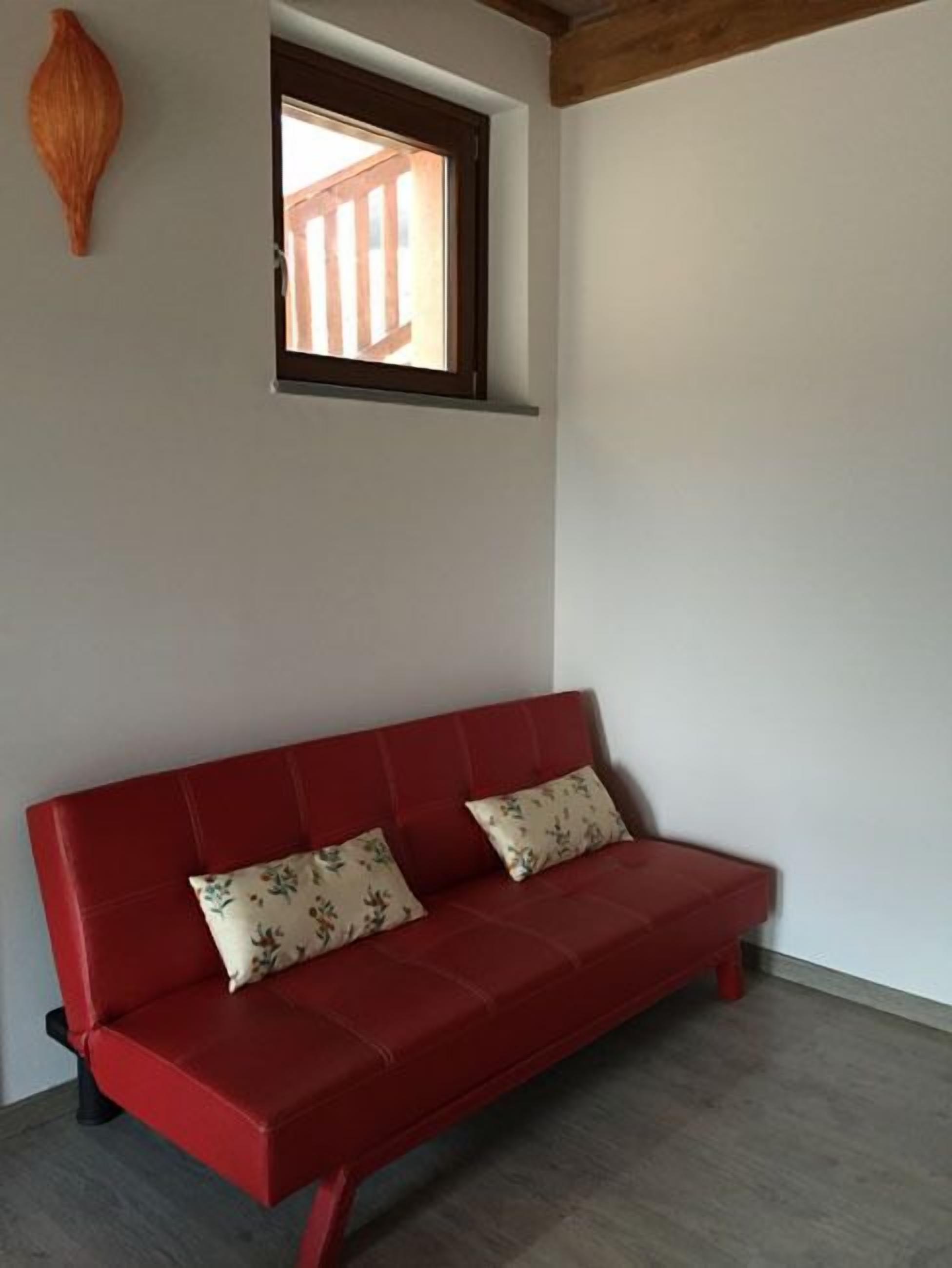 Un sofá rojo con cojines estampados en una habitación con una ventana pequeña y techo de vigas de madera.