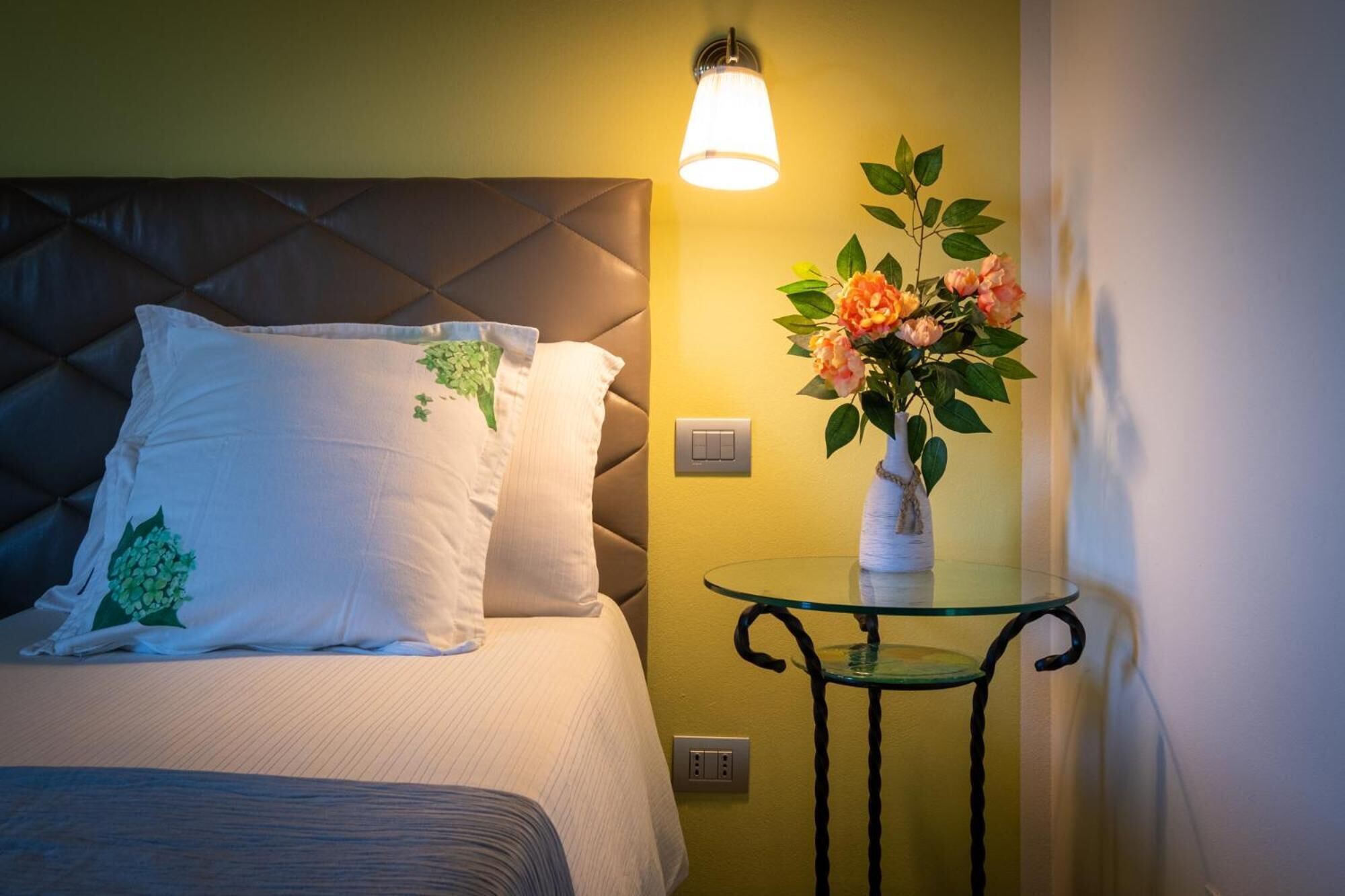 Una habitación de hotel con cama, una mesita de noche con un florero y una lámpara de pared.