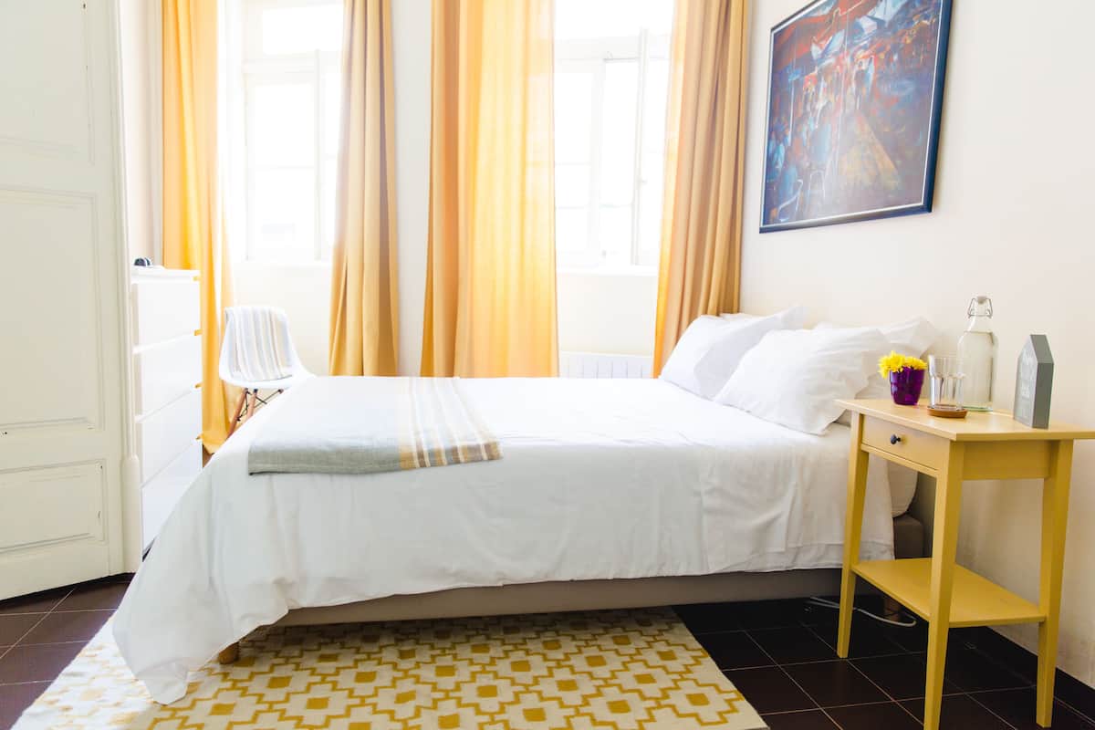 A hotel room with a bed, a bedside table, a chair, a window with curtains, and a painting on the wall.