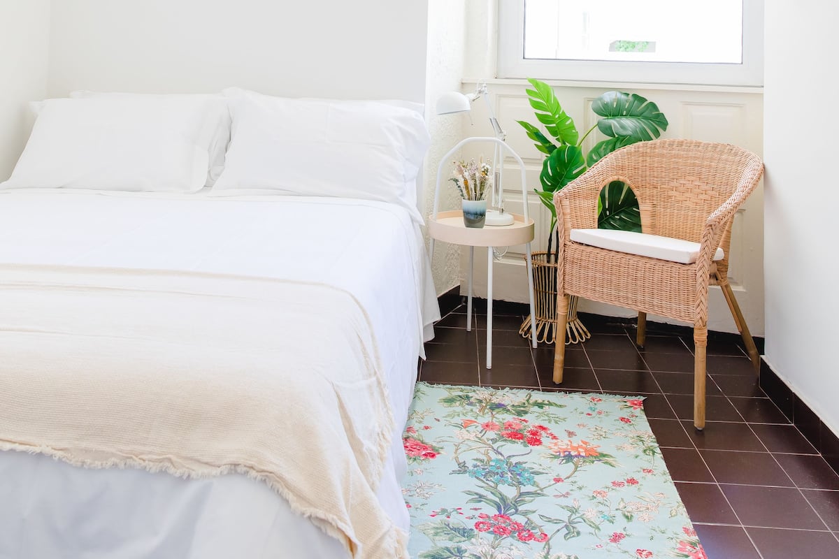 A bedroom with a bed, a window, a chair, and a small table with a lamp and a plant.