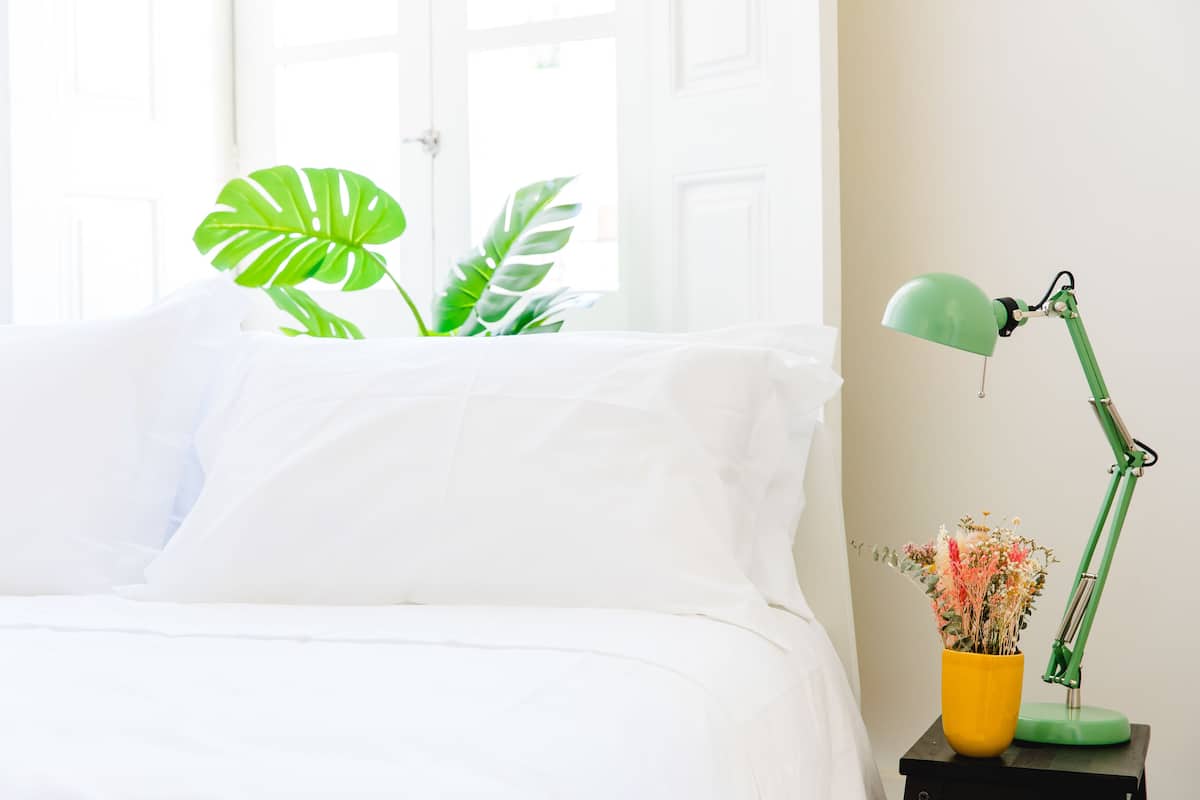 A neatly made bed with white linens, a green desk lamp, and a yellow vase with flowers on a bedside table.