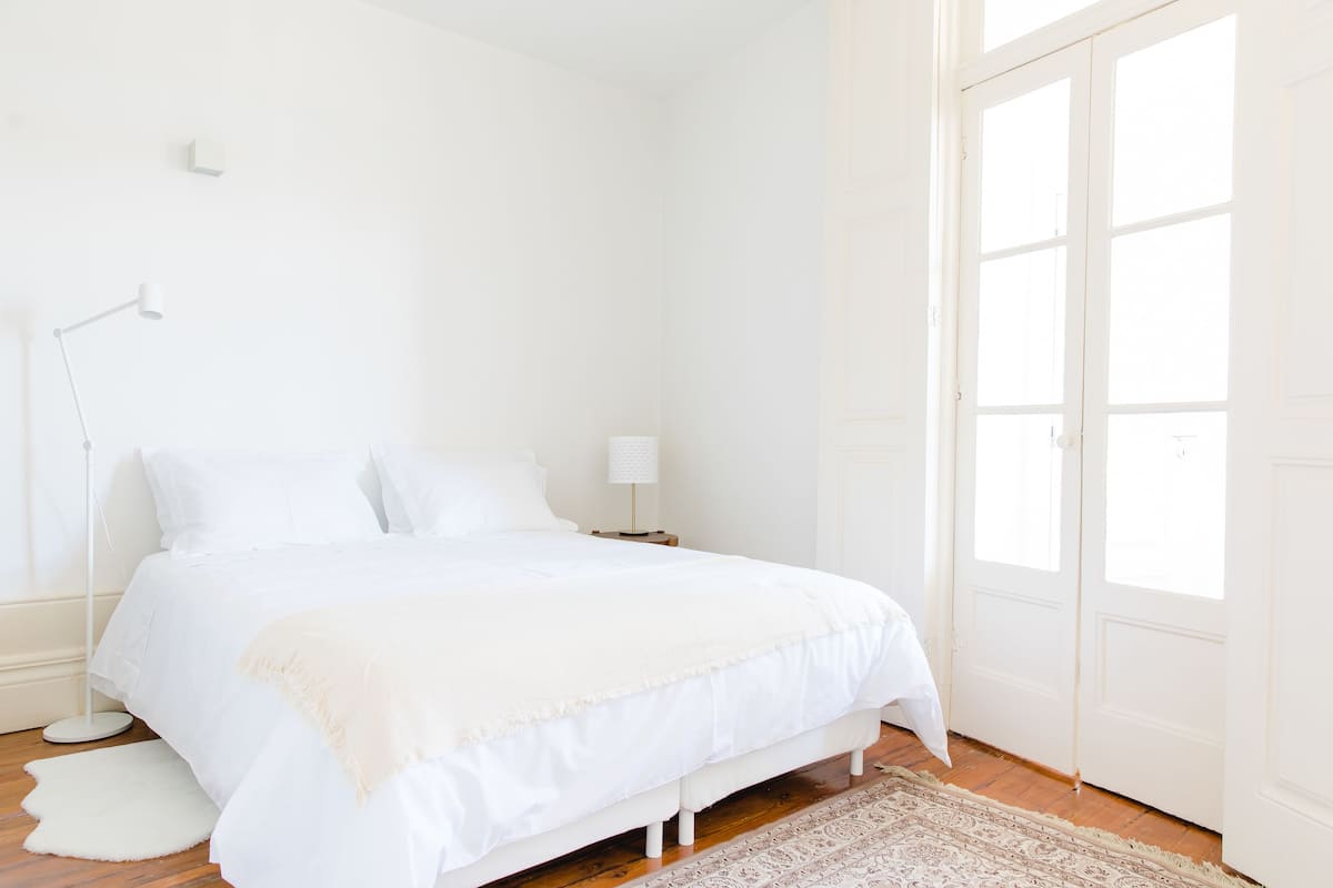 A minimalist bedroom with a large bed, a bedside table with a lamp, and a door leading to a bright room.