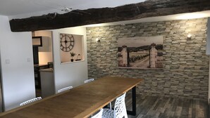 A dining area with a wooden table and white chairs, a stone wall with a vineyard painting, and a clock on the wall.