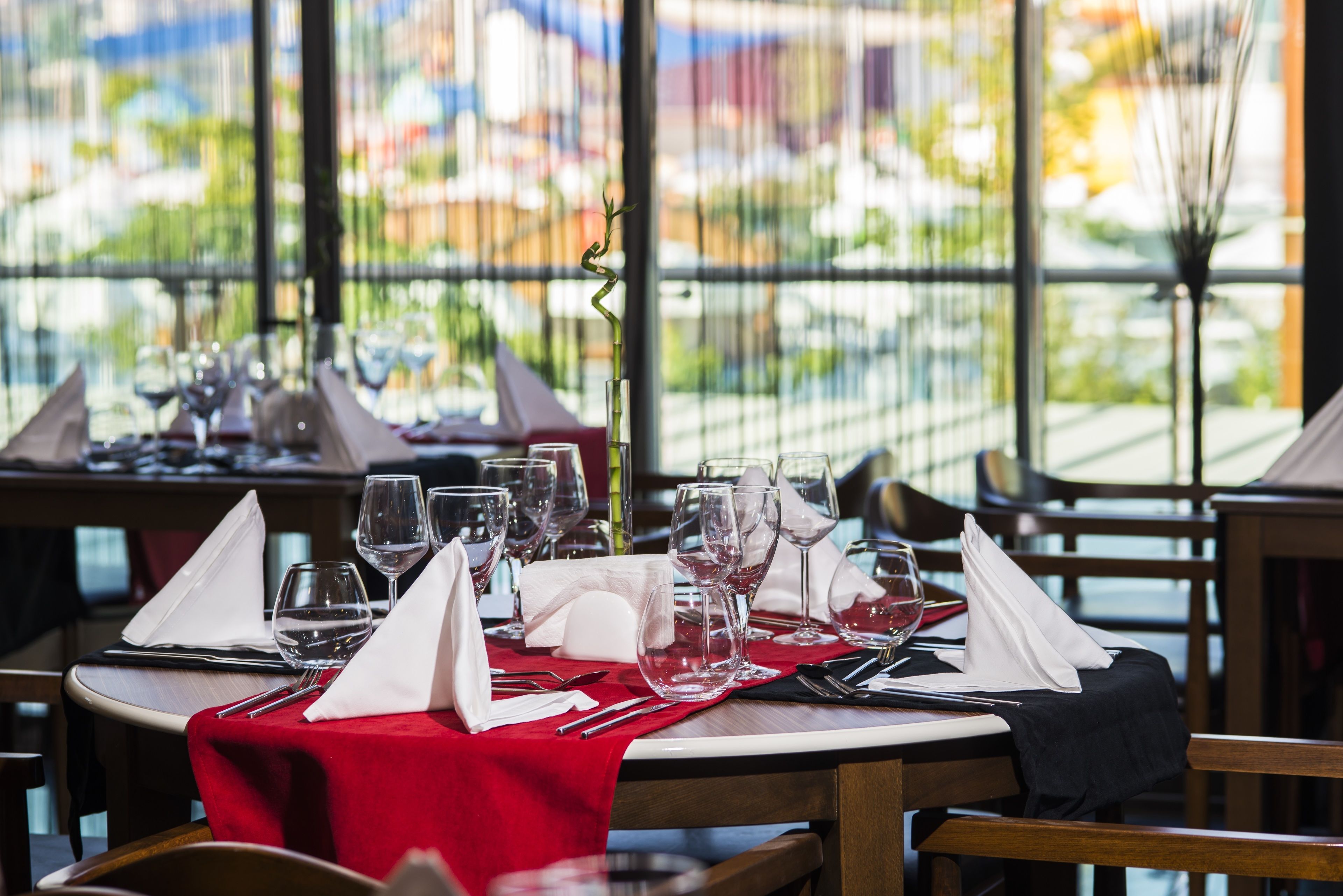 Mesa de restaurante preparada con copas de vino, cubiertos y servilletas, con vistas a un paisaje urbano a través de amplios ventanales.