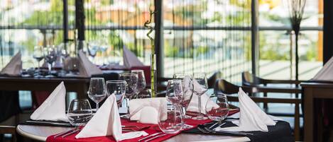 Mesa de restaurante preparada con copas de vino, cubiertos y servilletas, con vistas a un paisaje urbano a través de amplios ventanales.