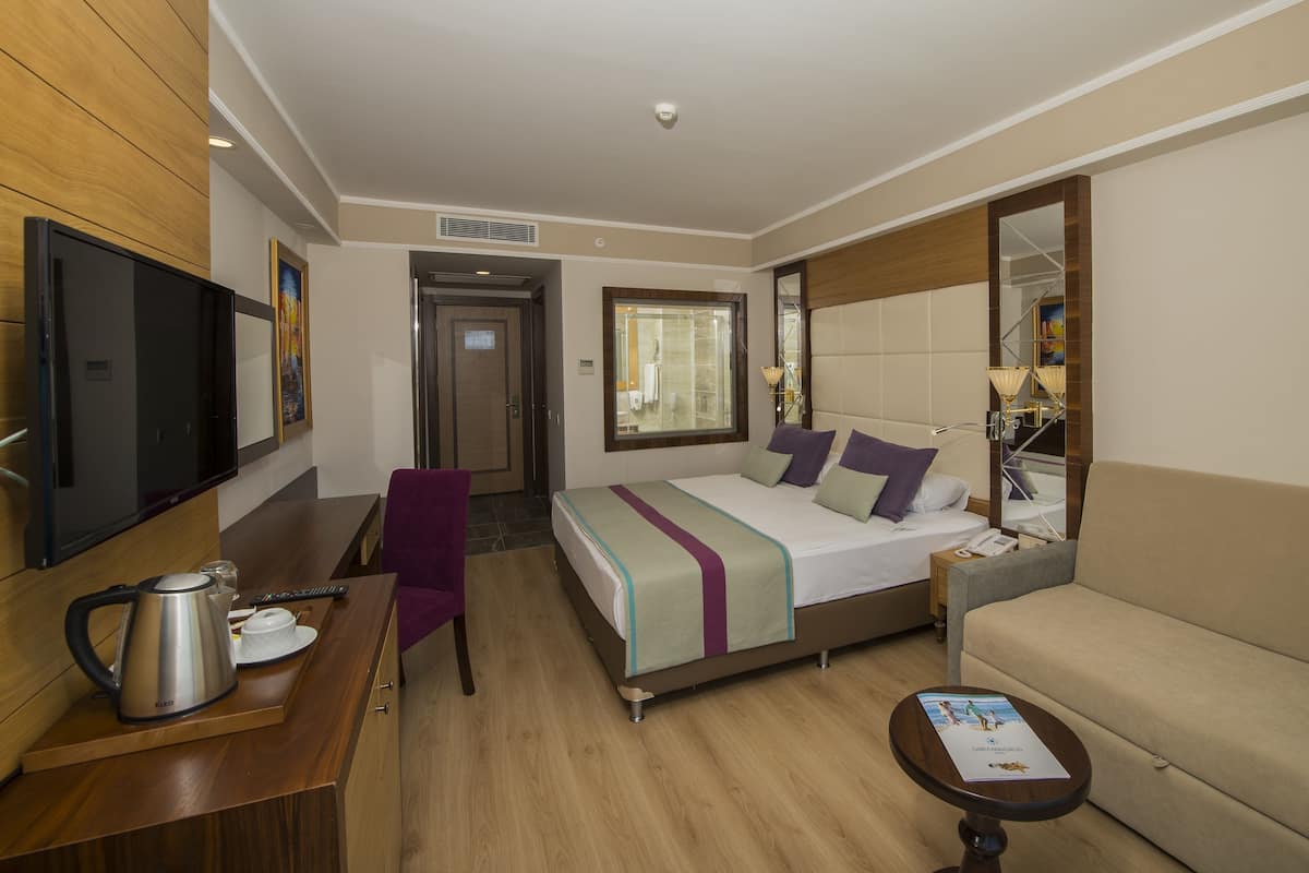 Habitación de hotel con cama, sofá, escritorio con hervidor, silla, televisor y ventana.
