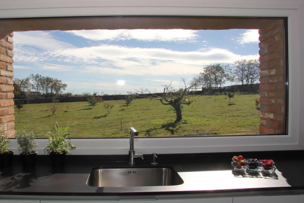 A kitchen with a large window overlooking a grassy field and trees.