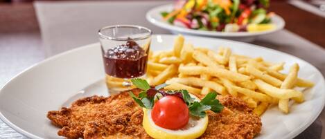A plate with a breaded cutlet, lemon slice, and cherry tomato garnish, accompanied by French fries and a small glass of sauce.