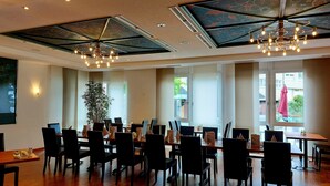 Un comedor amplio con varias mesas y sillas, una planta grande en maceta, y un diseño de techo moderno con iluminación empotrada.