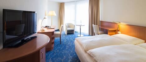 A hotel room with a flat-screen TV, a desk with a lamp, two chairs, a small round table, and a bed with white linens.