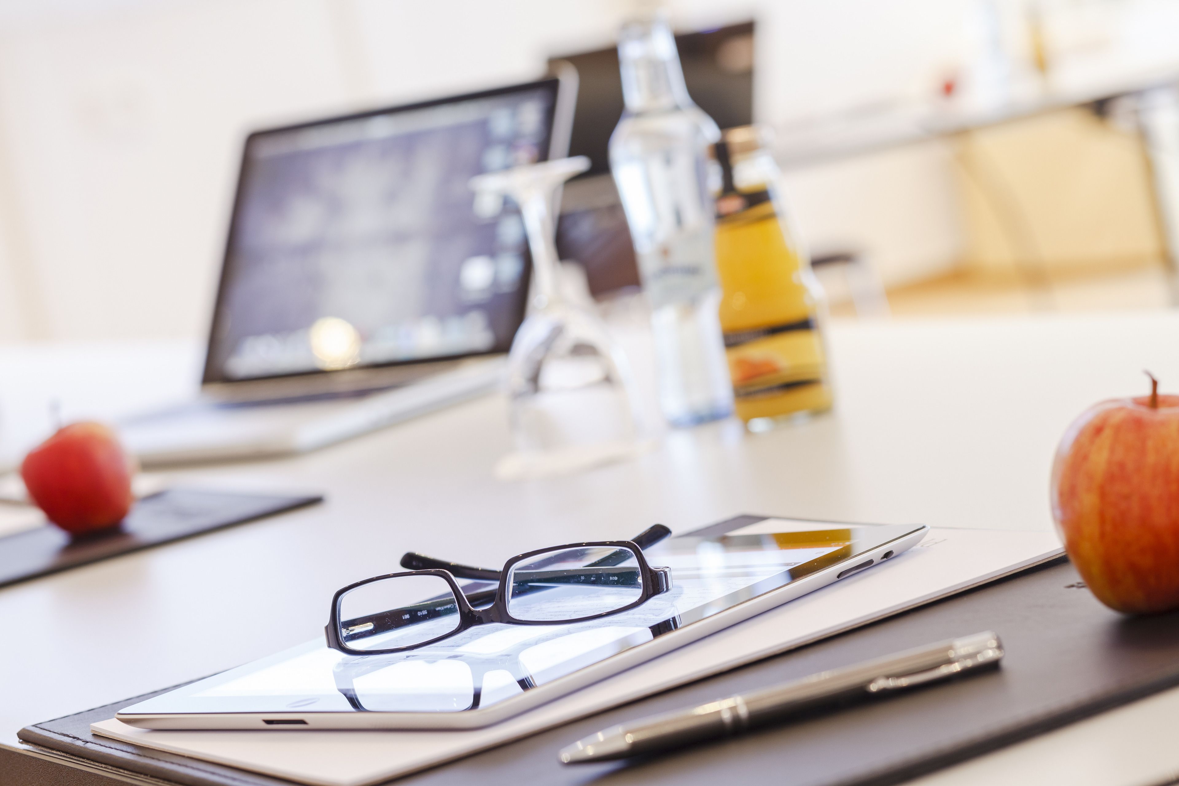 A desk with a tablet, glasses, a pen, and an apple.