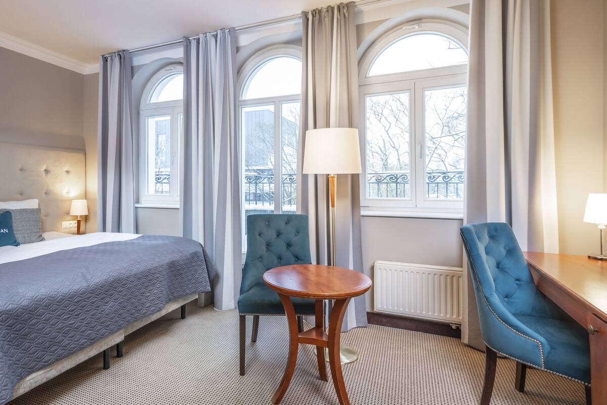 A hotel room with a bed, a chair, a table, and large windows with curtains.