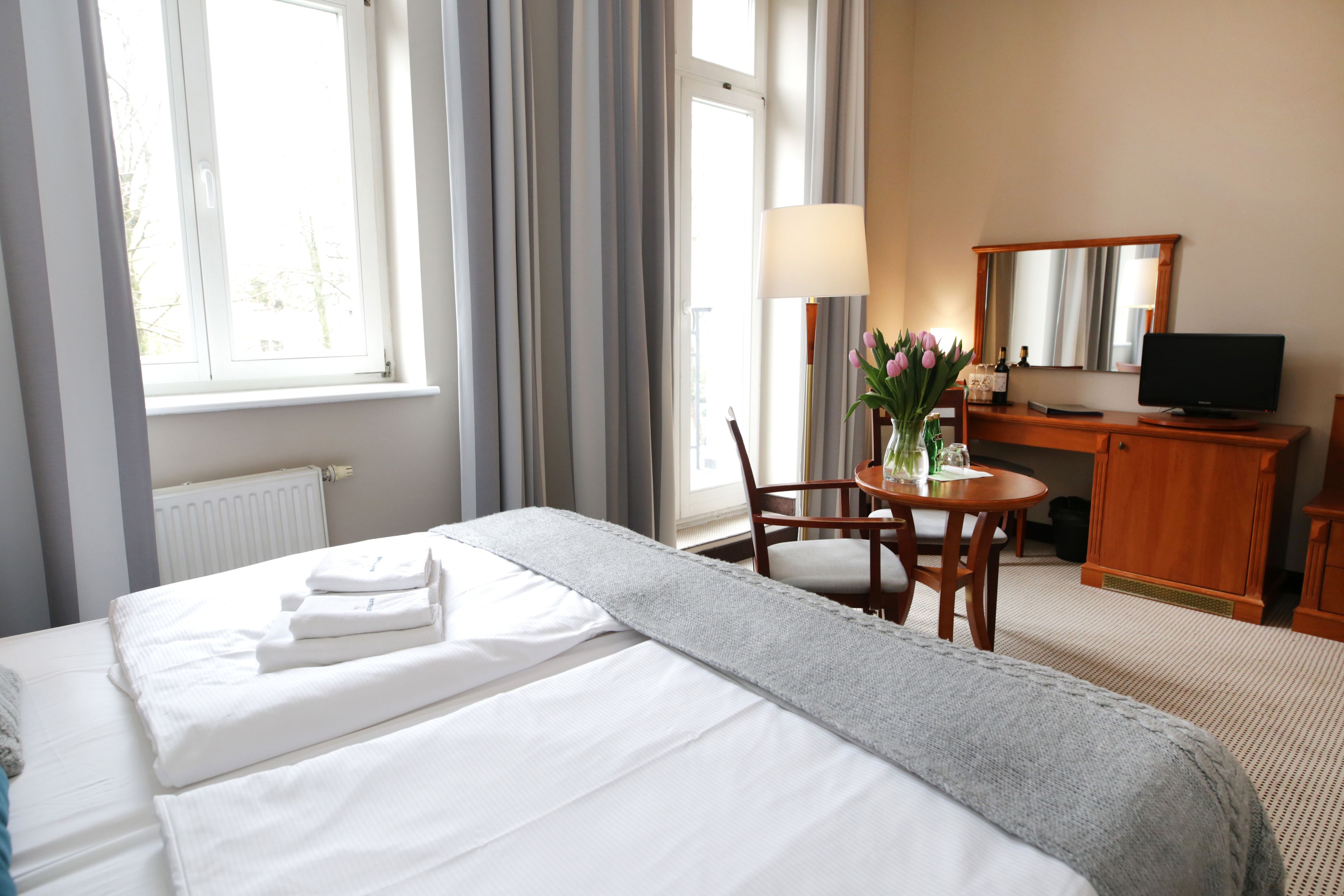 Habitación de hotel con cama, escritorio, silla, lámpara, televisor y ventana con cortinas.