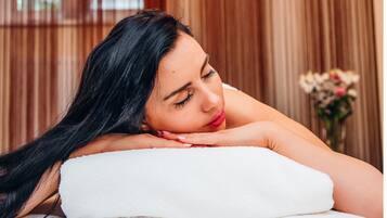 A person lying on a white massage table with a white towel, in a room with wooden paneling and a vase of flowers in the background.