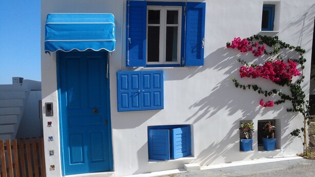 Ein Gebäude mit weißer Fassade und blauen Fensterläden, einer blauen Tür und einem kleinen Fenster mit rosa Blumen.