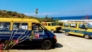 Un fourgon jaune et bleu, sur lequel on peut lire les inscriptions « Sarakino Studios » et « Gavdos », est garé sur une zone sablonneuse, offrant une vue sur la mer et les collines en arrière-plan.