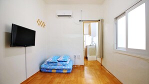 A hotel room with a wooden floor, a TV mounted on the wall, a bed with a blue floral bedspread, and a window with a view of the outside. - Dodam Guest House (Busan)