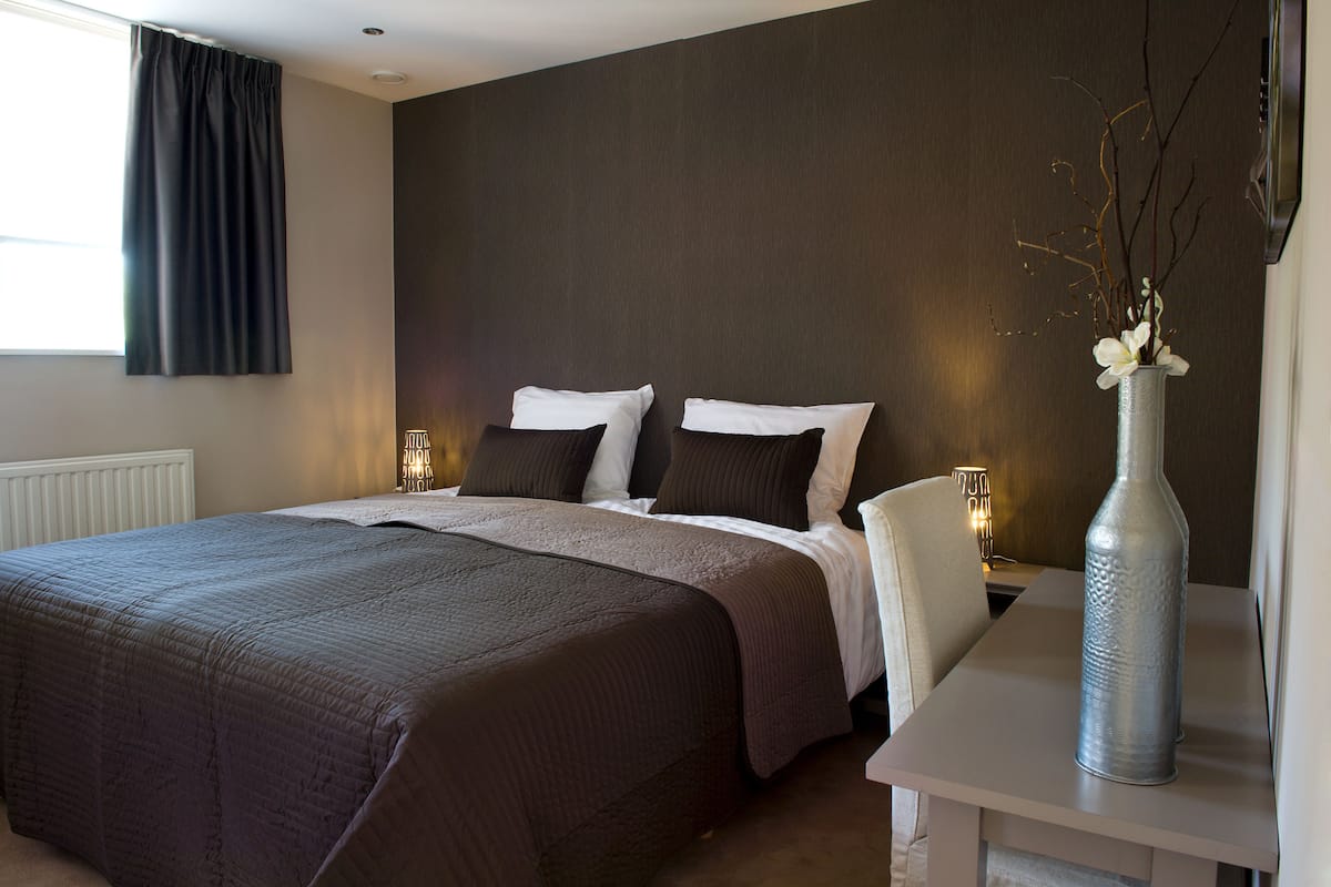 A hotel room with a large bed, two bedside tables, a nightstand, and a vase with branches.