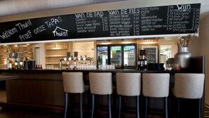 Zona de bar con un menú en pizarra que enumera diversas bebidas y tapas. Hay cuatro taburetes y una barra con botellas y vasos.