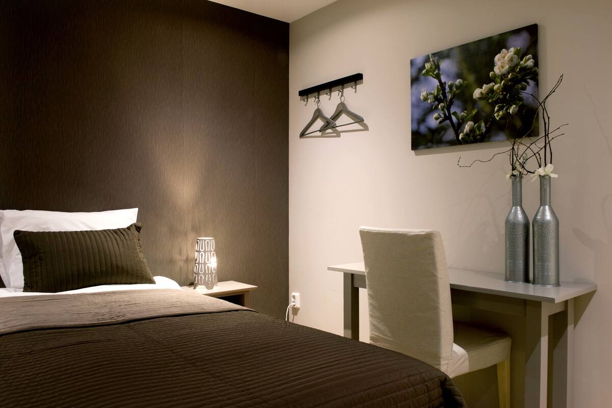 A hotel room with a bed, bedside table, chair, desk, and a painting on the wall.