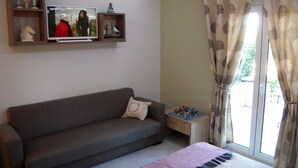 Une pièce avec un canapé gris, un téléviseur fixé au mur, une petite table ornée de figurines et une fenêtre avec des rideaux à motifs.