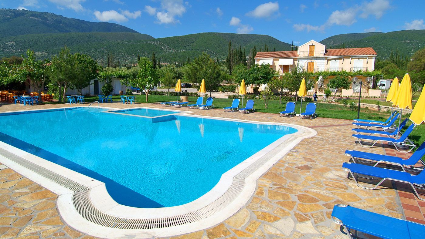 A swimming pool with blue water, surrounded by blue lounge chairs and yellow umbrellas, set against a backdrop of green hills and a building with a red roof.