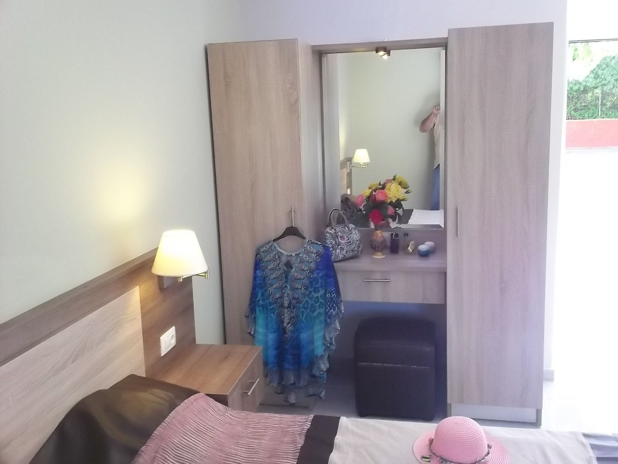 Una camera d'albergo con un letto, un comodino con una lampada, una cassettiera con uno specchio e un appendiabiti.