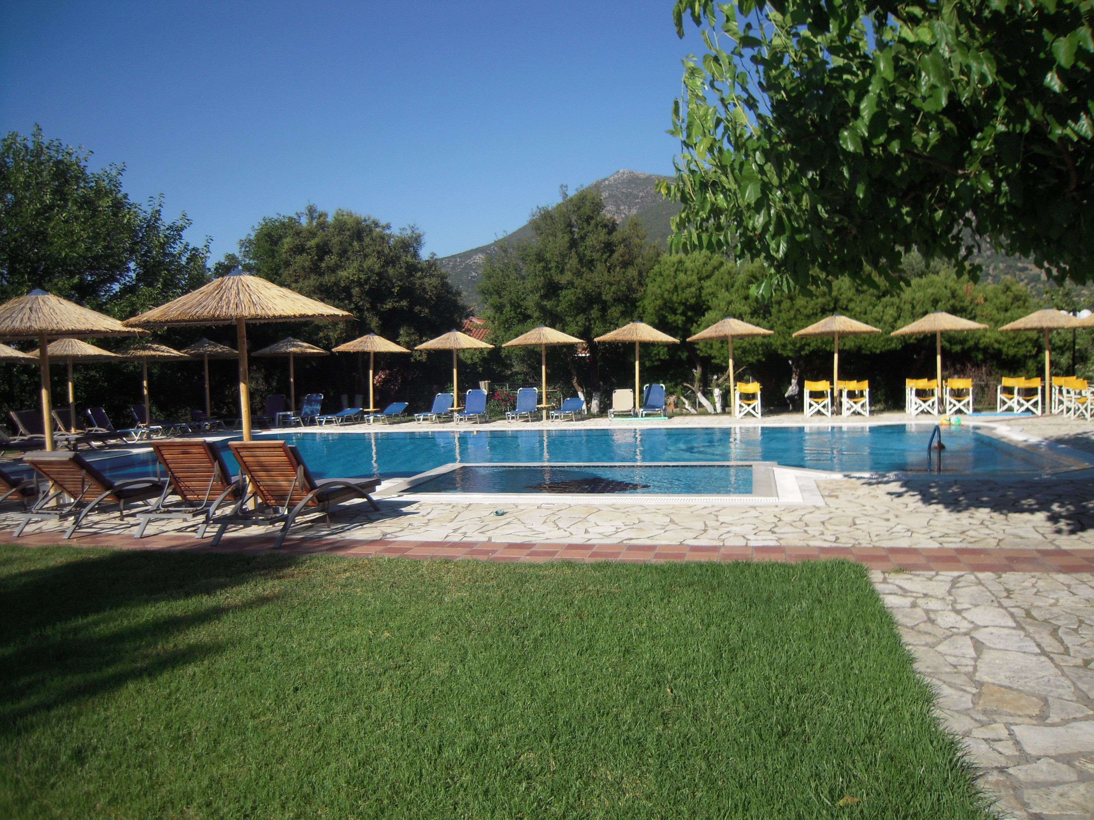 Ein Poolbereich mit Liegestühlen und Sonnenschirmen, umgeben von Grünflächen und einem Berg im Hintergrund.