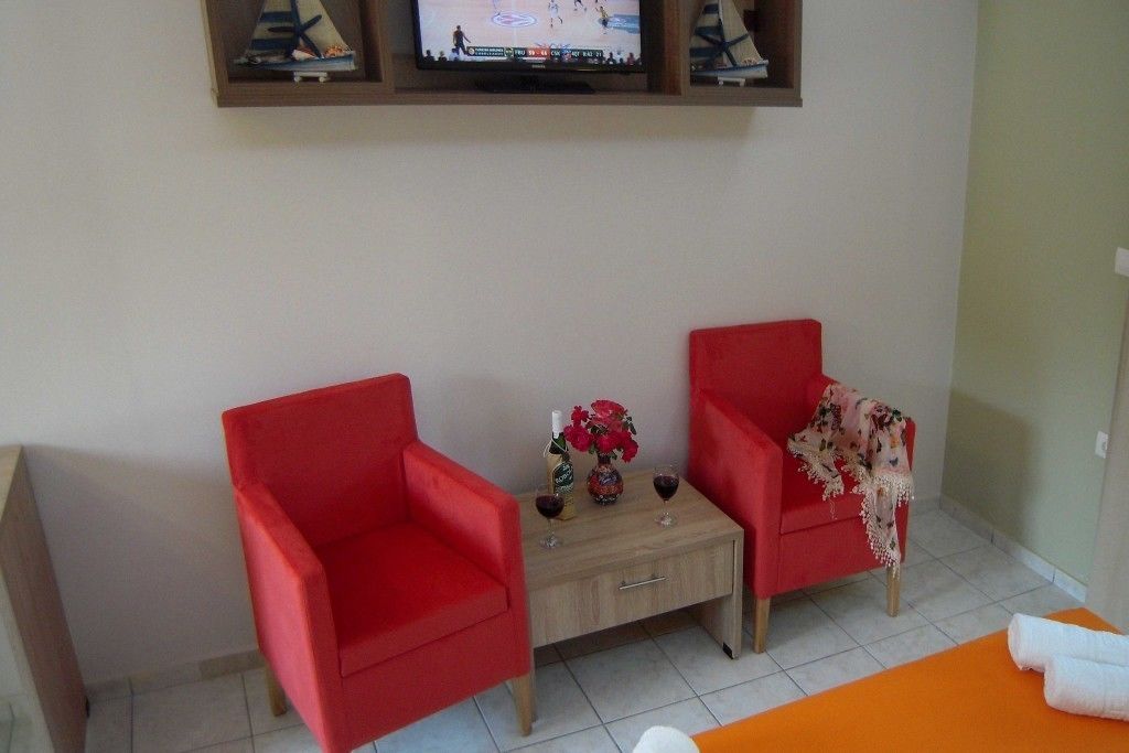 A room with two red chairs, a small wooden table with a vase and wine glasses, and a television mounted on the wall.