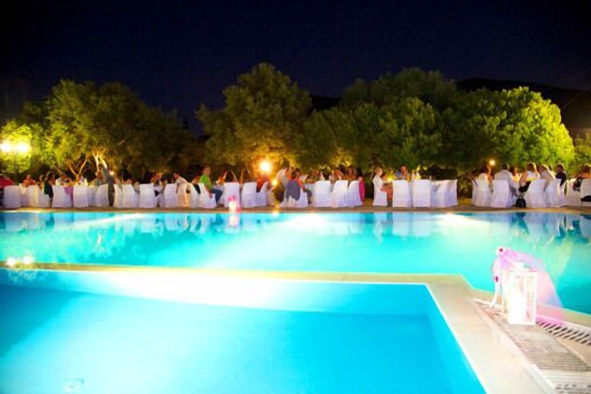 A poolside gathering with people seated on white chairs, surrounded by trees and a pool.