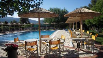 A poolside area with lounge chairs and tables, umbrellas, and a view of mountains in the background.