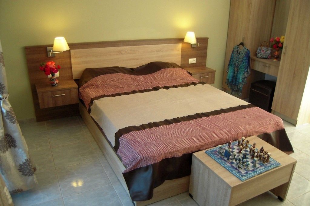 Une chambre à coucher avec un lit, deux tables de chevet avec des lampes, une petite table avec un vase de fleurs, une tête de lit en bois et une petite table sur laquelle est posé un jeu d’échecs.