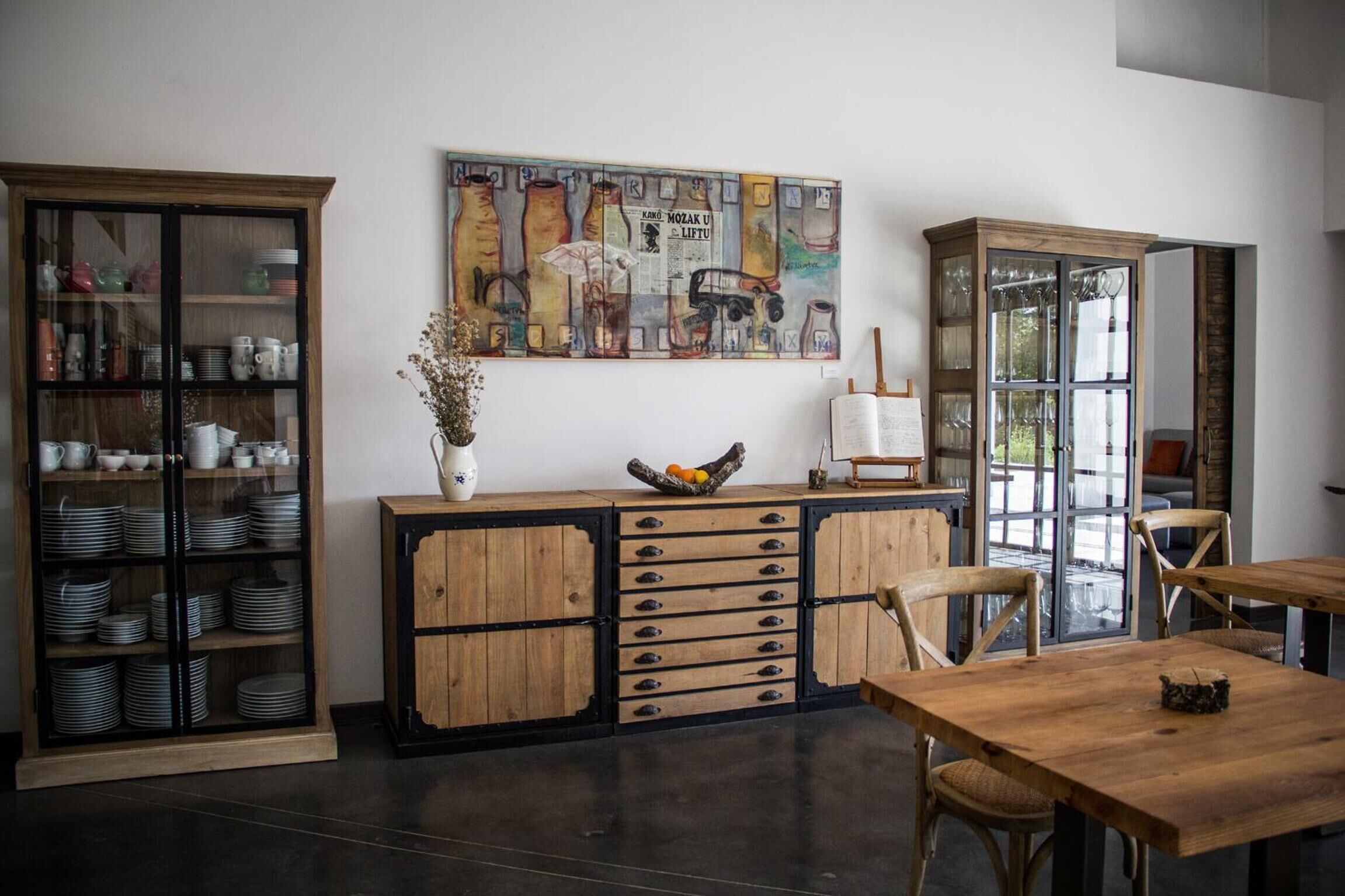 A dining area with a wooden table and chairs, a cabinet with glass doors displaying dishes, and a wall-mounted painting.